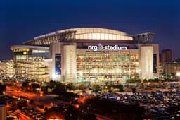 Houston — NRG Stadium: stadium lit up at dusk with city lights and surrounding parking in the foreground.