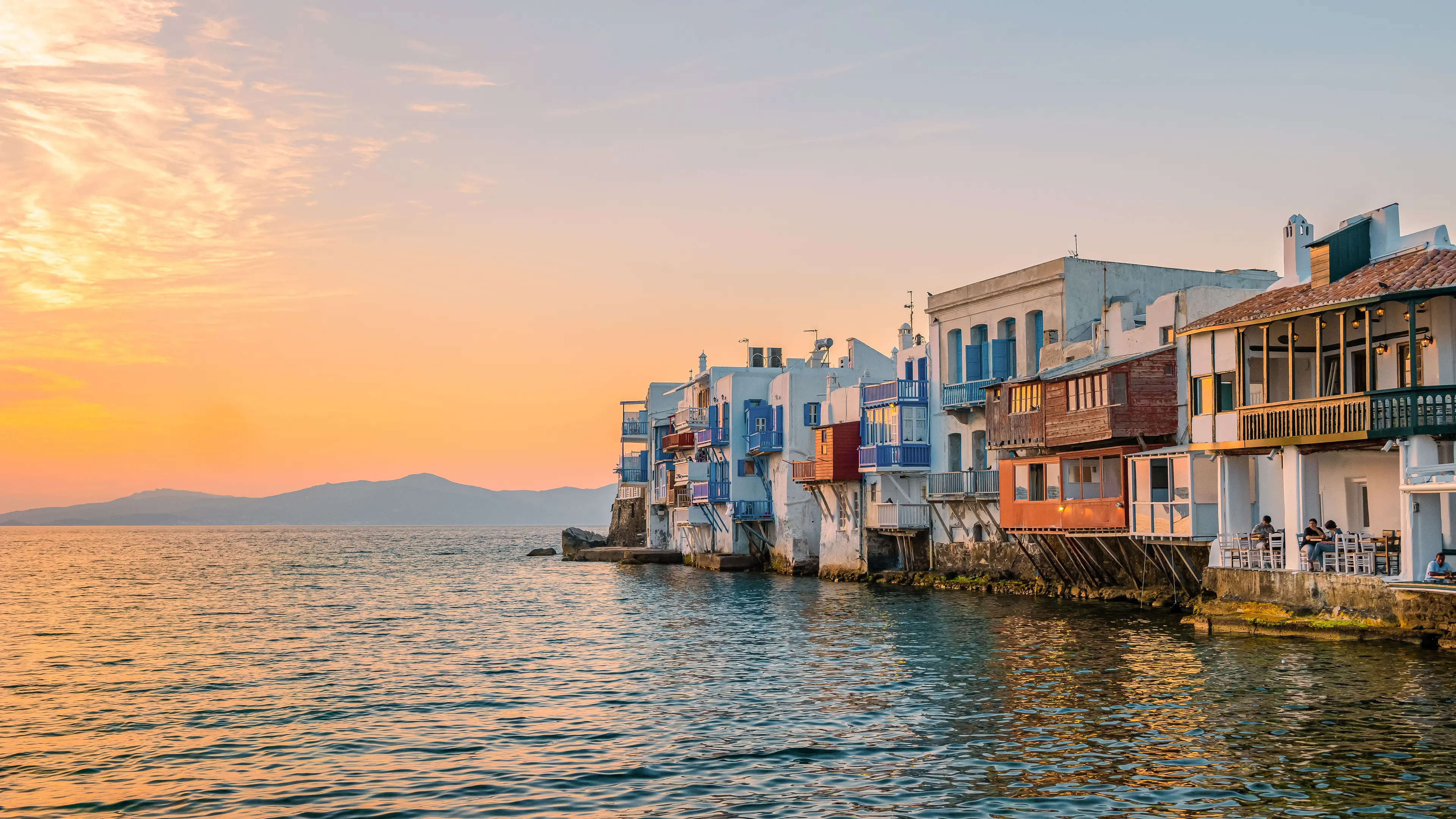Colorful waterfront buildings in Little Venice, Mykonos, at sunset, with reflections on the Aegean Sea.