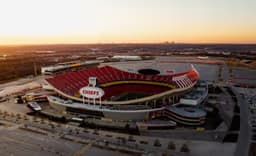 Kansas City — Arrowhead Stadium: aerial view of a stadium with red seating at sunset, set among open fields.