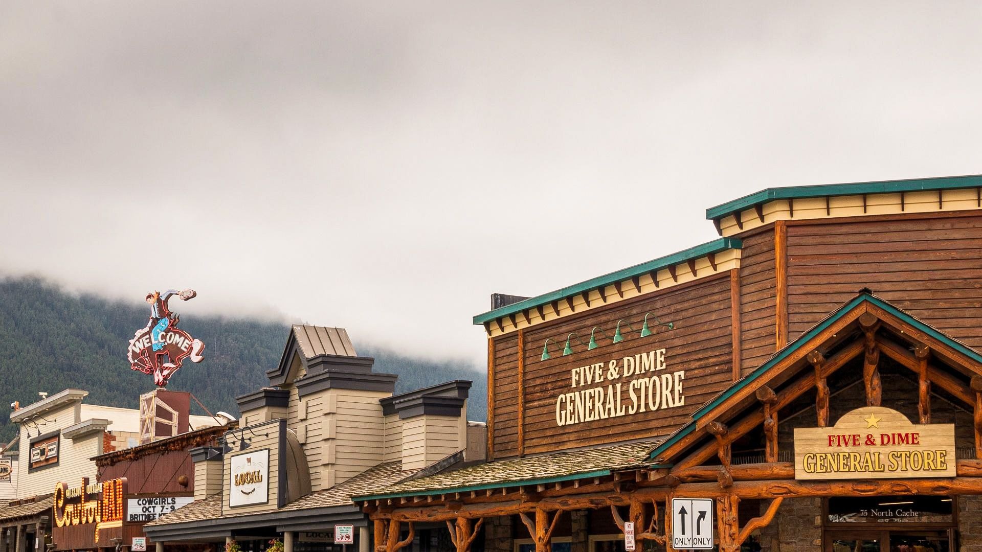 A row of Western-inspired storefronts in downtown Jackson, Wyoming, featuring the Five & Dime General Store and the iconic cowboy ‘Welcome’ sign against a cloudy mountain backdrop.