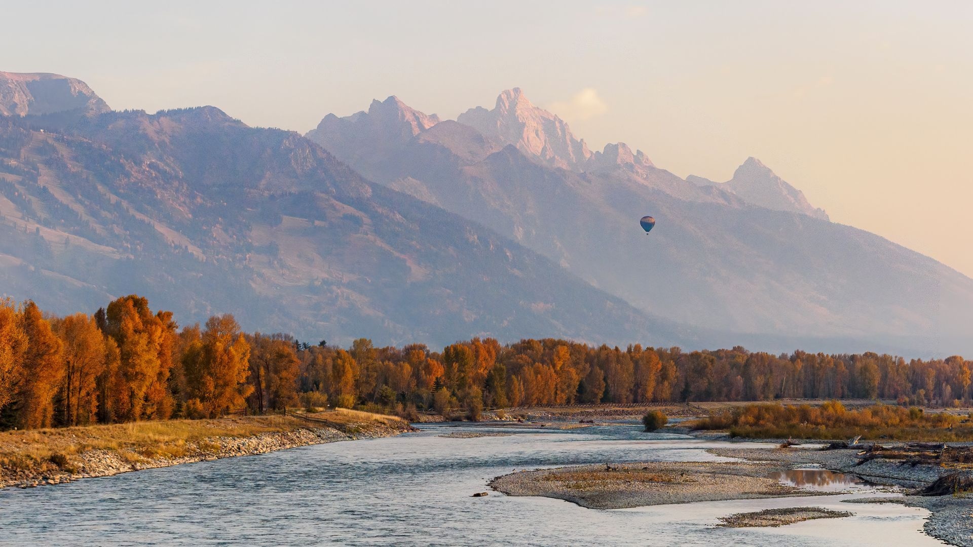 A hot air balloon drifts above the Snake River with autumn-colored trees along the banks and the jagged peaks of the Teton Range rising in the background at sunset.