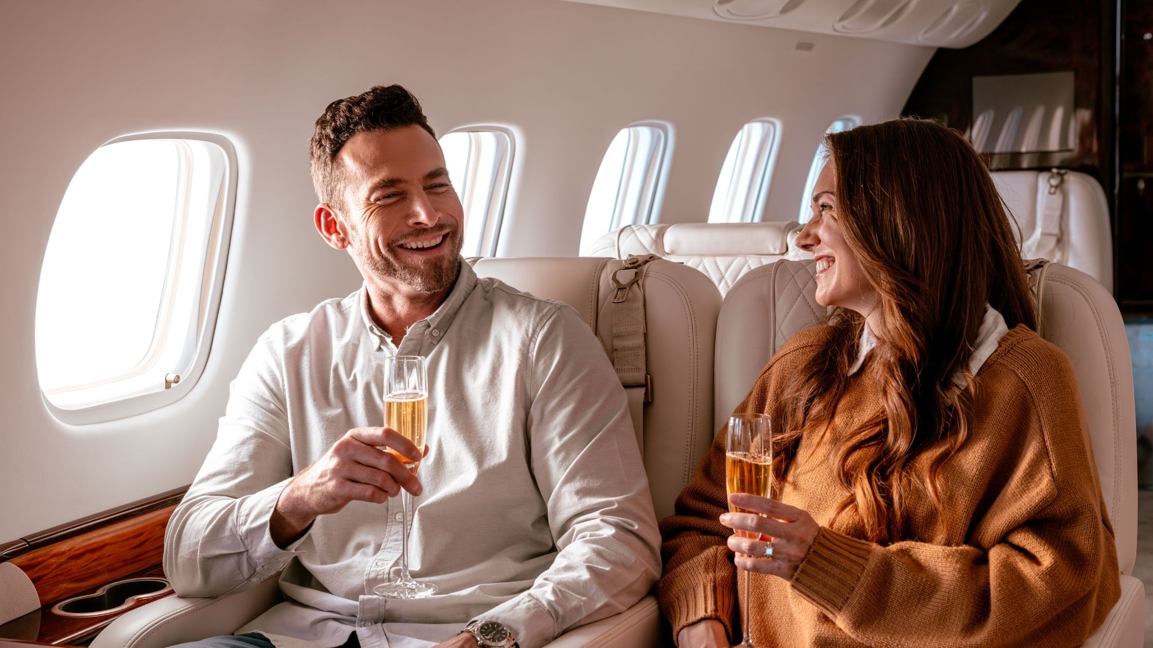 Smiling couple enjoying champagne aboard a private jet, seated in luxury leather seats with natural light streaming through the windows