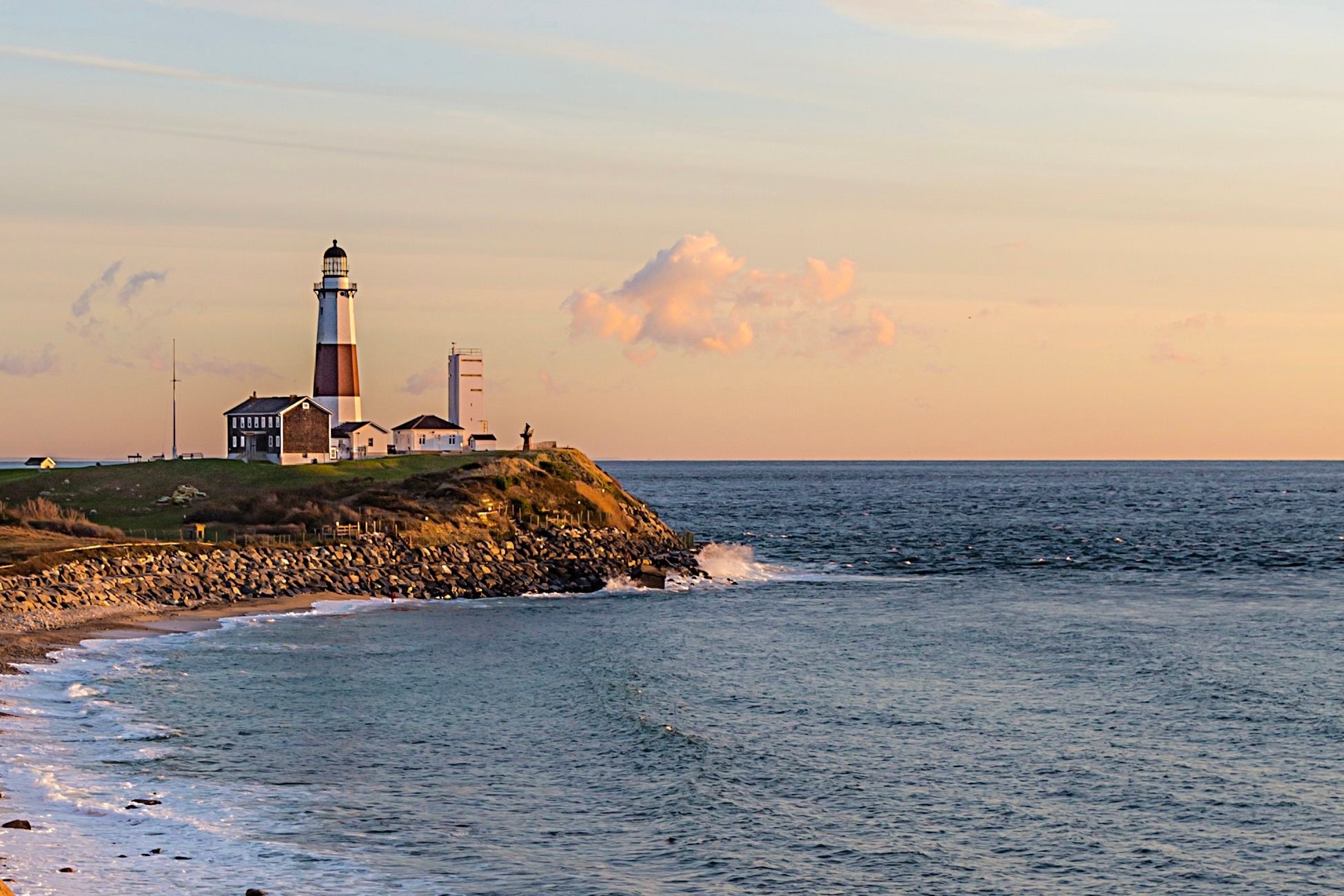 View of Montauk Point Lighthouse perched on a rocky cliff overlooking the Atlantic Ocean at sunset, with gentle waves and a soft pastel sky in the background.