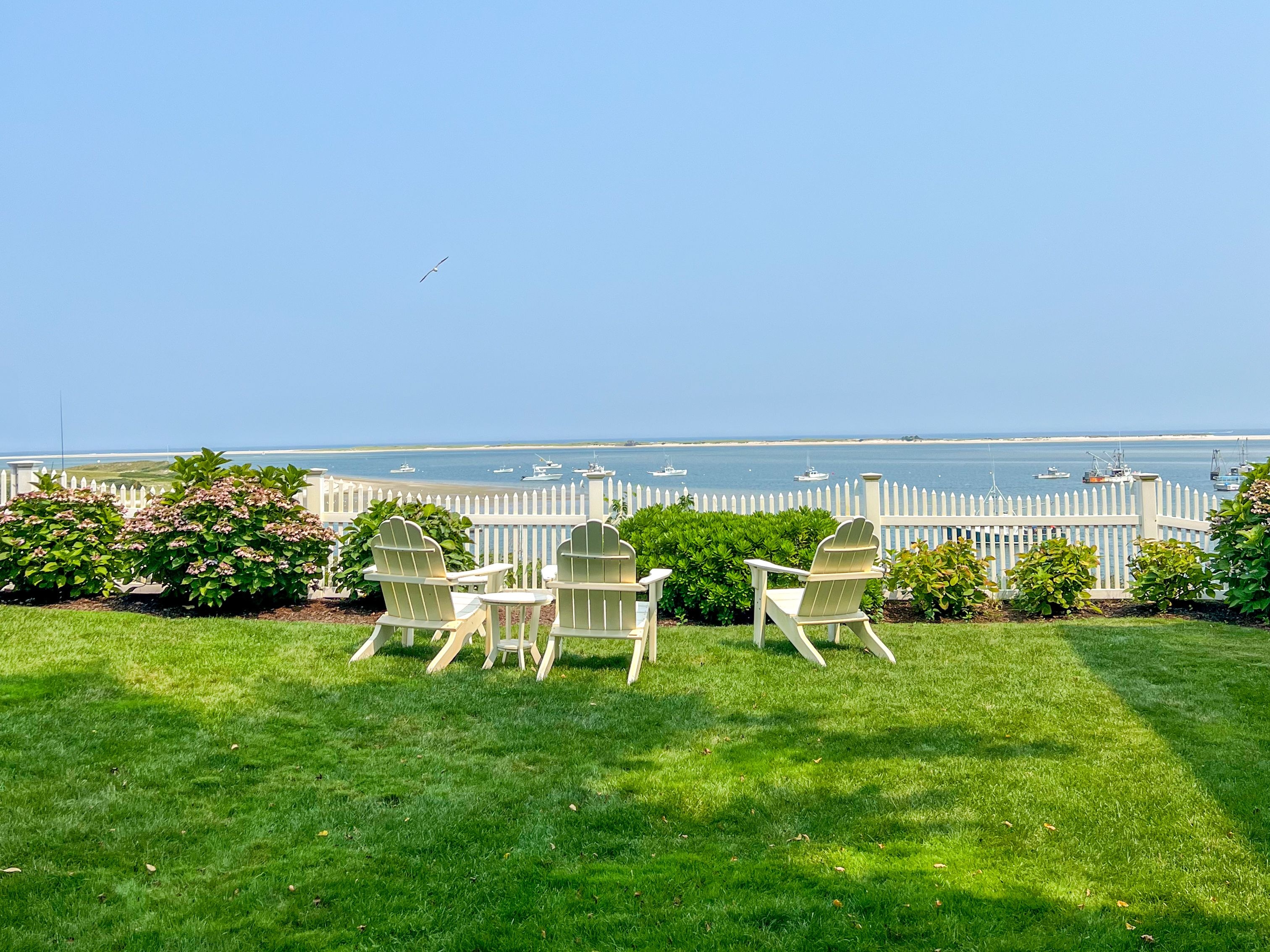 Three white Adirondack chairs on a manicured lawn facing a white picket fence with a view of boats on the water in the Hamptons, surrounded by blooming flowers under a clear blue sky.